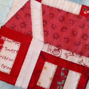 May include: A red and white patchwork quilt block with a house design. The house has a white roof and red walls. The quilt block has a white background with a red border. The text "Joyful Bells, Sweet Warm Hugs Grow" is written on the house.