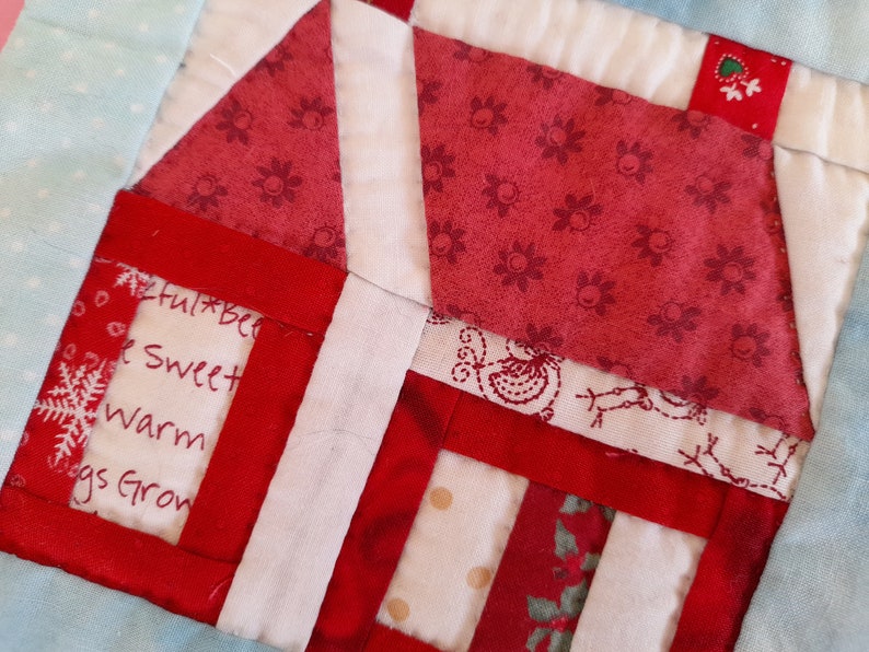 May include: A red and white patchwork quilt block with a house design. The house has a white roof and red walls. The quilt block has a white background with a red border. The text "Joyful Bells, Sweet Warm Hugs Grow" is written on the house.