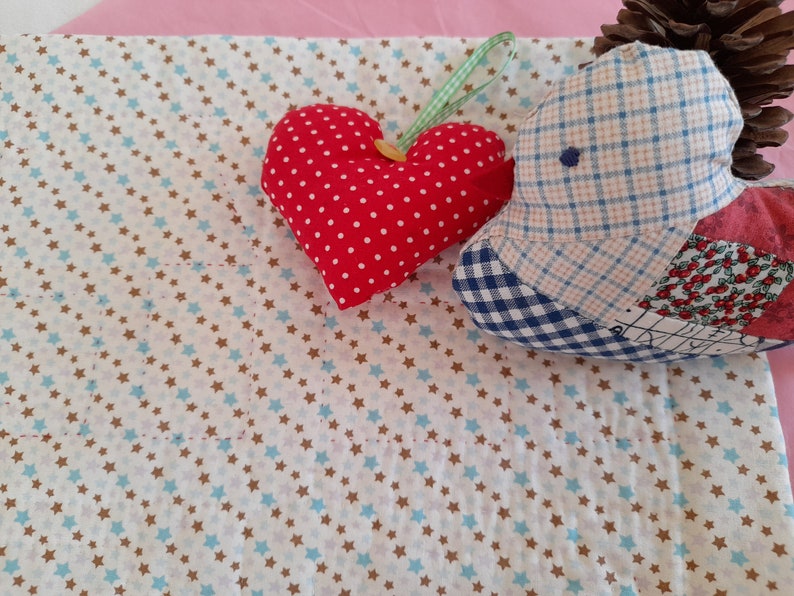 May include: A white fabric with a pattern of small blue and brown stars. A red heart-shaped pillow with white polka dots and a blue and white plaid pillow are on the fabric.