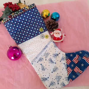 Può includere: Una calza di Natale patriottica con un motivo a stelle blu e bianco, un design a bandiera americana e un motivo ad albero. La calza è decorata con ornamenti, una statuina di Babbo Natale e bacche. La calza è su una superficie rosa.