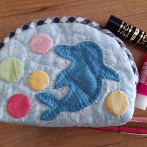 Può includere: Una pochette in tessuto trapuntato blu con un design a delfino e pois colorati. La pochette ha un bordo a quadri bianco e nero.