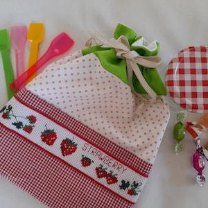Peut inclure: Un sac à cordon en tissu vichy blanc et rouge avec un dessus en tissu vert. Le sac a un motif à pois rouge et blanc et un motif brodé de fraises avec le mot "STRAWBERRY". Le sac est rempli de petites cuillères en plastique et de bonbons.
