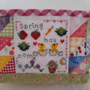 Può includere: Una pochette in tessuto patchwork rosa e verde con un ricamo a punto croce bianco con due pulcini gialli, fragole, una carota e il testo "Spring has come".