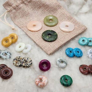 May include: A collection of circular stone beads with center holes in various colors including green, blue, white, brown, and spotted patterns, arranged around a tan burlap drawstring pouch on a textured white surface.