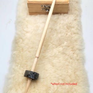May include: A light-colored wooden drop spindle featuring a tapered shaft and a dark gray, textured circular whorl with a speckled pattern. The text *whorl not included is printed in red near the bottom of the spindle shaft.