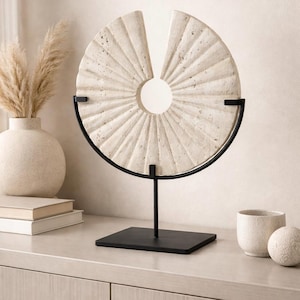 May include: A beige, circular stone sculpture with a radial pattern, mounted on a black metal stand. The sculpture has a central hole and a section removed. A vase with dried grass, books, a small cup, and a sphere are also visible.