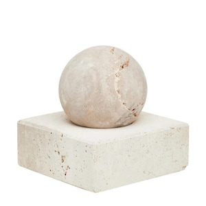 May include: A light beige, spherical sculpture sits on a square, light beige base. The sphere has a textured surface with visible grain and a crack. The base is a solid, square block with a slightly rough texture.