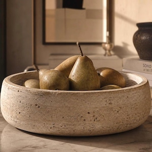 May include: A round, textured, beige stone bowl filled with several green and brown pears. The bowl sits on a marble surface. The pears have brown stems and are various sizes.