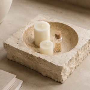May include: A square, beige travertine stone bowl with a rough, natural exterior and a smooth, circular interior basin. Inside the bowl are two cylindrical white wax candles of varying heights and a small glass jar filled with wooden matches.