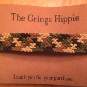 May include: A camouflage-patterned friendship bracelet made with woven string. The bracelet is displayed on a brown card with the text "The Gringa Hippie" and "Thank you for your purchase."