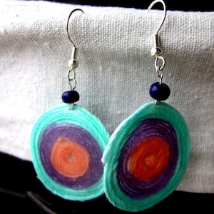Puede incluir: Un par de pendientes circulares turquesa, morado y naranja con ganchos plateados y cuentas azul oscuro. Los pendientes están hechos de papel.