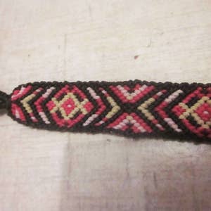 Puede incluir: Un brazalete de amistad negro con un patrón geométrico en rosa, blanco y beige.