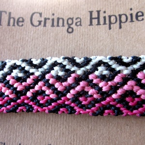 May include: A close-up of a pink, black, and white friendship bracelet with a chevron pattern. The bracelet is on a brown background with the text "The Gringa Hippie" printed on it.