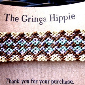 May include: A close-up of a woven fabric band with a chevron pattern in brown, beige, light blue, and purple. The band is on a brown card with the text "The Gringa Hippie" and "Thank you for your purchase."
