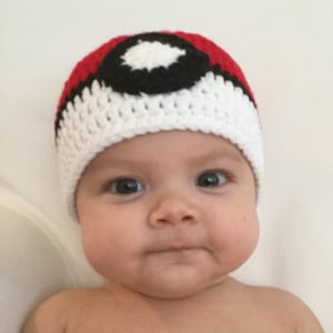 May include: A baby wearing a white and red crocheted hat with a black and white Pokeball design.