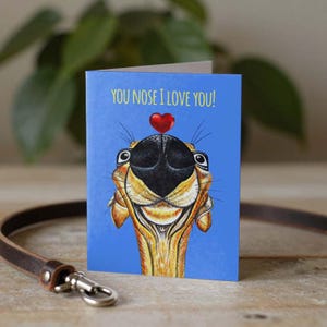 May include: A blue greeting card with a cartoon dog face and a red heart above its nose. The text "YOU NOSE I LOVE YOU!" is printed at the top. A brown leather leash with a silver clasp is in the foreground. The card is on a wooden surface.