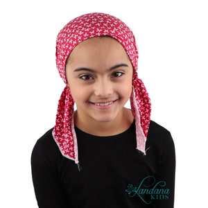 May include: A red and white patterned head covering with ties, worn by a young person. The head covering is made of a soft fabric and features a repeating cross-like design. The person is wearing a black long-sleeved shirt. The brand name "Landana KIDS" is visible.