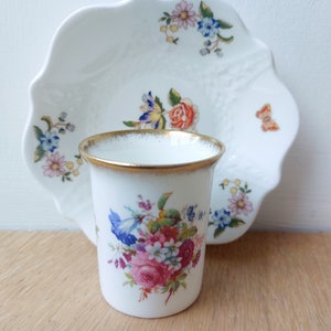 May include: A white porcelain teacup and saucer set with a floral pattern. The cup has a gold rim and the saucer has a scalloped edge.