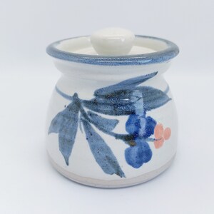 May include: A white ceramic jar with a blue and pink floral design and a white lid. The jar has a blue rim and a white base.