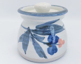 May include: A white ceramic jar with a blue and pink floral design and a white lid. The jar has a blue rim and a white base.