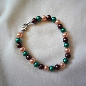 May include: A beaded bracelet featuring alternating colors of pink, green, and brown beads. The bracelet is secured with a silver-colored clasp. The beads are round and evenly spaced, creating a simple yet elegant design.