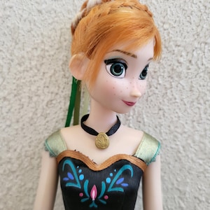 Puede incluir: Mu&ntilde;eca con cabello rojo peinado en trenza, ojos verdes y pecas. La mu&ntilde;eca viste un vestido negro con detalles florales turquesa y rosa y un collar con colgante dorado. El vestido de la mu&ntilde;eca tiene ribetes dorados y detalles verdes.