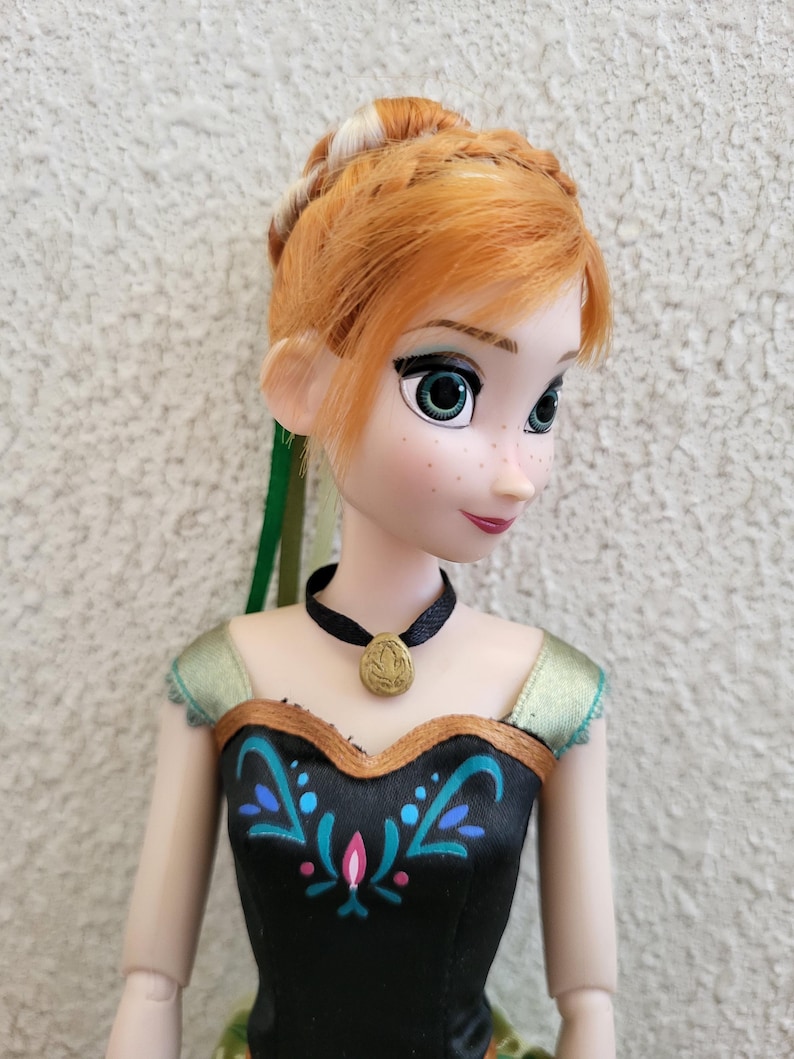 Puede incluir: Mu&ntilde;eca con cabello rojo peinado en trenza, ojos verdes y pecas. La mu&ntilde;eca viste un vestido negro con detalles florales turquesa y rosa y un collar con colgante dorado. El vestido de la mu&ntilde;eca tiene ribetes dorados y detalles verdes.
