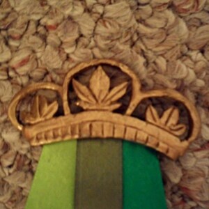 Puede incluir: Una corona dorada con tres dise&ntilde;os en forma de hoja. La corona se asienta sobre una cinta con rayas verticales verdes, verde oliva y verde oscuro. El fondo es una superficie texturizada de color marr&oacute;n claro.