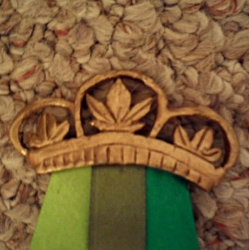 Puede incluir: Una corona dorada con tres dise&ntilde;os en forma de hoja. La corona se asienta sobre una cinta con rayas verticales verdes, verde oliva y verde oscuro. El fondo es una superficie texturizada de color marr&oacute;n claro.