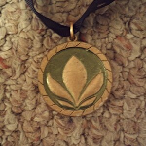 Puede incluir: Un colgante redondo con un marco dorado y un centro verde oscuro. El centro presenta un dise&ntilde;o floral dorado. El colgante cuelga de una cinta negra.