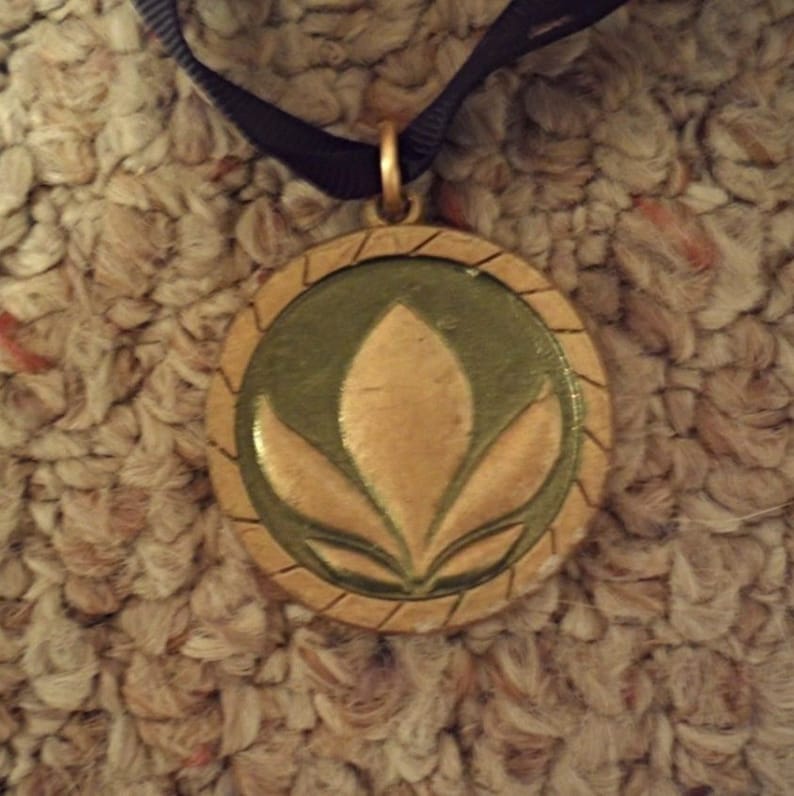 Puede incluir: Un colgante redondo con un marco dorado y un centro verde oscuro. El centro presenta un dise&ntilde;o floral dorado. El colgante cuelga de una cinta negra.