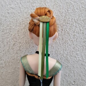 Puede incluir: Mu&ntilde;eca con cabello rojo peinado en un mo&ntilde;o, adornada con un accesorio para el cabello de color dorado y tres cintas en tonos verdes y crema. La mu&ntilde;eca lleva una parte superior negra con detalles verdes y dorados.