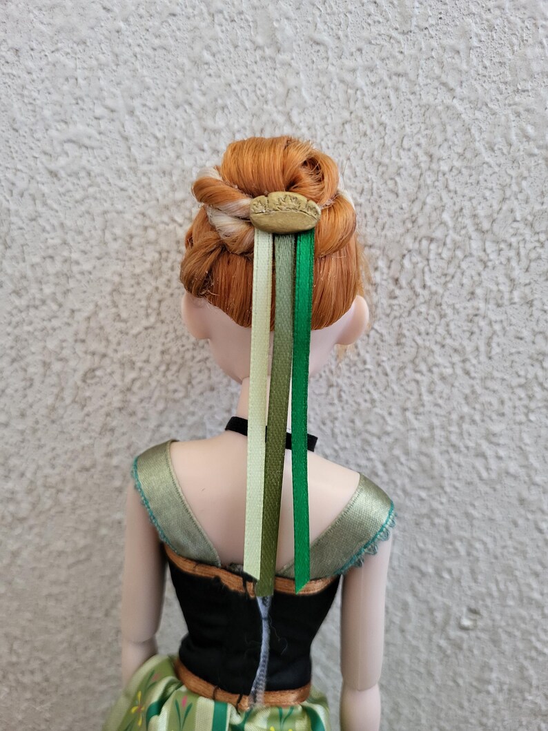 Puede incluir: Mu&ntilde;eca con cabello rojo peinado en un mo&ntilde;o, adornada con un accesorio para el cabello de color dorado y tres cintas en tonos verdes y crema. La mu&ntilde;eca lleva una parte superior negra con detalles verdes y dorados.