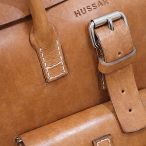 May include: Close-up of a tan leather bag with the embossed word "HUSSAR". The bag features a handle, a flap pocket, and a silver buckle closure. White stitching accents the leather.