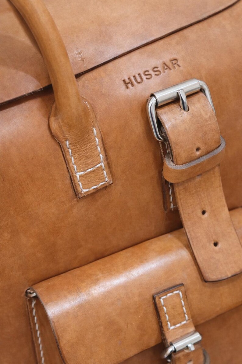 May include: Close-up of a tan leather bag with the embossed word "HUSSAR". The bag features a handle, a flap pocket, and a silver buckle closure. White stitching accents the leather.