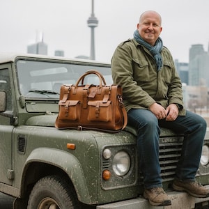 May include: A tan leather briefcase with multiple pockets and a top handle rests on the bonnet of a green vintage SUV. The briefcase has a classic design and is made of high-quality leather.