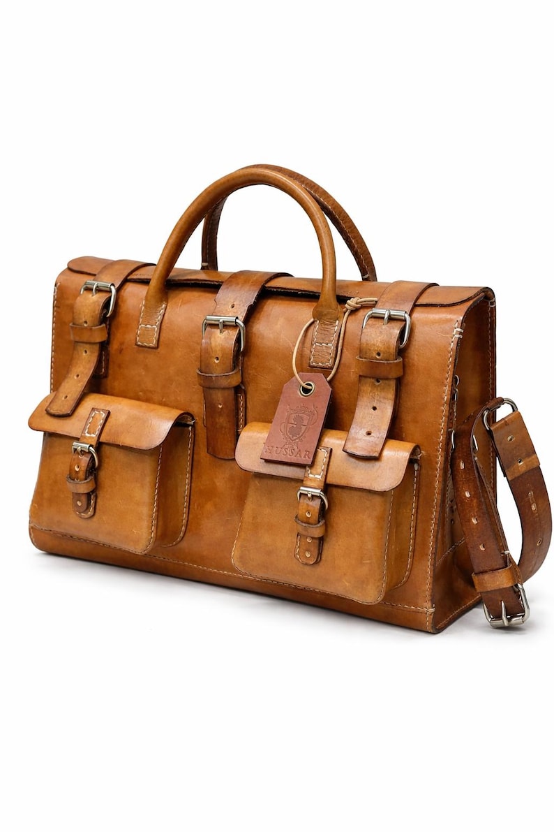 May include: A tan leather briefcase with multiple pockets and straps. The bag has a top handle, a shoulder strap, and several buckles. A small tag with the word "RUSSARI" is attached to the front.