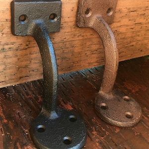 Puede incluir: Dos soportes de estante de hierro fundido, uno negro y otro marr&oacute;n, con un dise&ntilde;o curvo y una base redonda. Los soportes est&aacute;n montados en una superficie de madera.