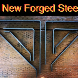 May include: Set of five black metal shelf brackets, in various sizes, arranged in a triangular pattern on a wooden surface. The brackets are made of forged steel and have a rustic, industrial look.