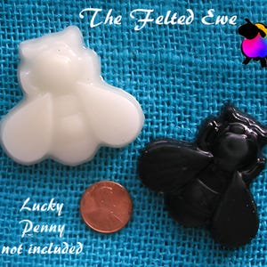 May include: Two felt bee ornaments, one white and one black, on a blue burlap background. The text "The Felted Ewe" is visible above the ornaments. A penny is visible below the ornaments.