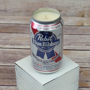 May include: A silver can candle with a Pabst Blue Ribbon beer label. The candle is on a white wooden block.