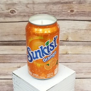 May include: Orange-scented candle in a can shaped like a Sunkist soda can. The can is orange with a blue Sunkist logo and the words "Orange Soda" and "12 fl oz (355 ml)".