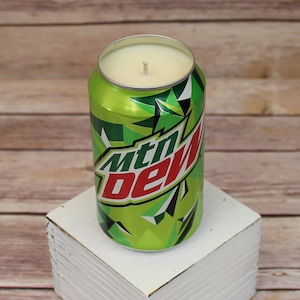 May include: A green and red Mountain Dew can with a white wax candle inside. The can is sitting on a white wooden surface.