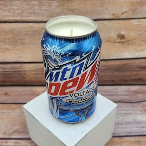 May include: A blue and white can candle with a white wax top and a wick. The can has the words "Mountain Dew Voltage" and "DEW Charged with Raspberry Citrus Flavor and Caffeine with Other Natural Flavors" printed on it.