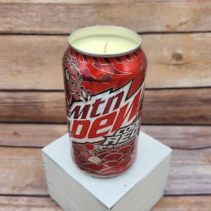 May include: A red and white can candle shaped like a Mountain Dew Code Red can. The candle has a yellow wax top and a wick.