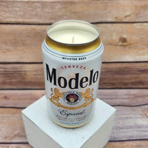 May include: A white and gold can candle shaped like a Modelo beer can. The candle has a white wax top and a gold label with the words "Modelo" and "Cerveza" printed on it. The label also features a crest with the words "Cerveceria Modelo Mexico 1925" and the text "Especial.  Brewed in Mexico."