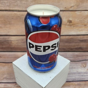 May include: A blue and red can candle with the Pepsi logo and the words "Wild Cherry" printed on the side. The candle is sitting on a white wooden box.