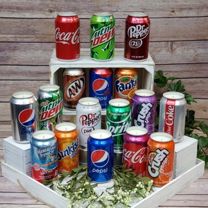 May include: A collection of 15 soda can-shaped candles in various colors and designs, including Coca-Cola, Pepsi, Dr. Pepper, Mountain Dew, Fanta, Crush, and Sprite. The candles are arranged on a white wooden shelf and tray.