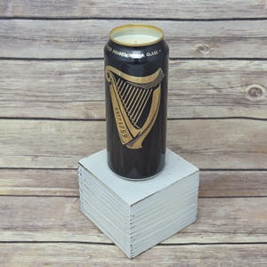 May include: A black candle in a Guinness can with a gold harp logo and the text "ESTD 1759". The candle sits on a small white square block against a wooden background.