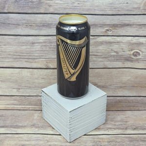 May include: A black and gold Guinness-branded candle in a can shape, with the harp logo and "ESTD 1759" printed on the front. The candle sits on a white square block against a wooden background.